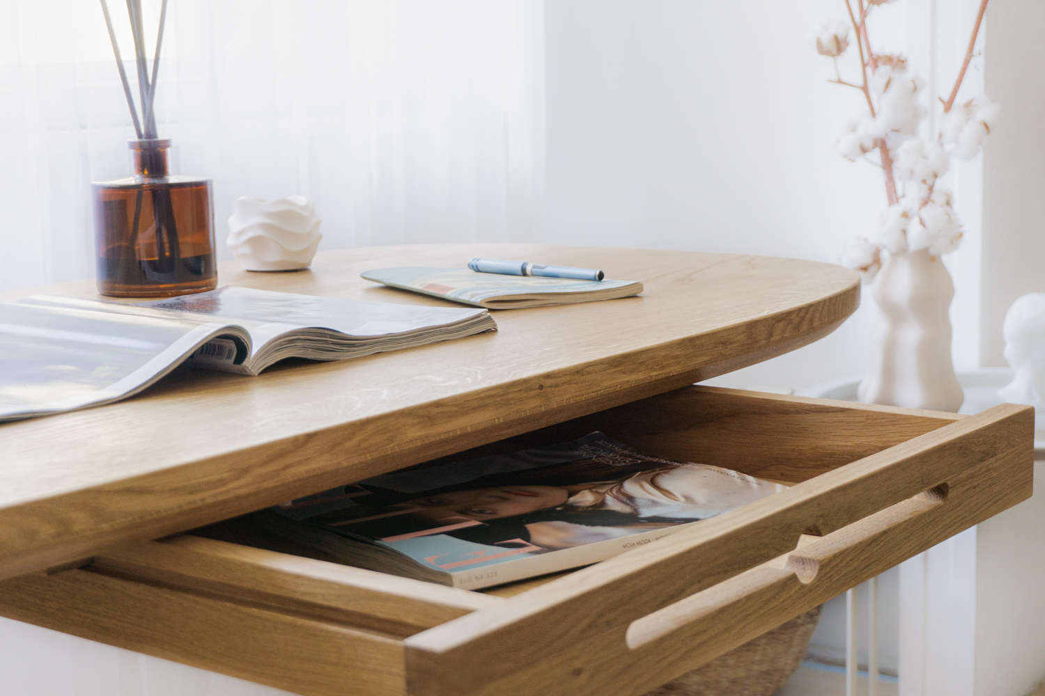 Close-up of a small writing desk with a drawer, made of solid oak and featuring a natural wood finish, in a home office next to a large window with a curtain