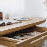 Close-up of a small writing desk with a drawer, made of solid oak and featuring a natural wood finish, in a home office next to a large window with a curtain