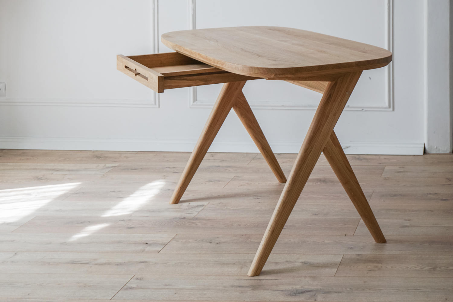Kunsst real wood vintage desk with drawer placed in a bright living room with warm oak flooring. The white wall behind features  elegant moulding. 