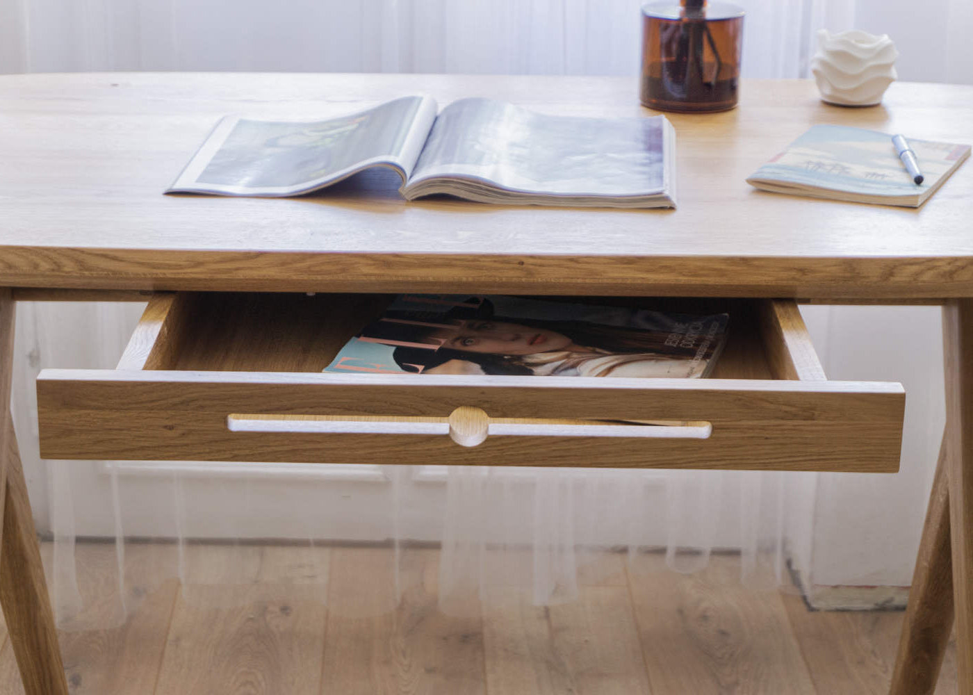 Solid wood vintage desk with drawer with an open book on oak desk top and a magazine inside a drawer, on a light wooden floor standing in front of window
