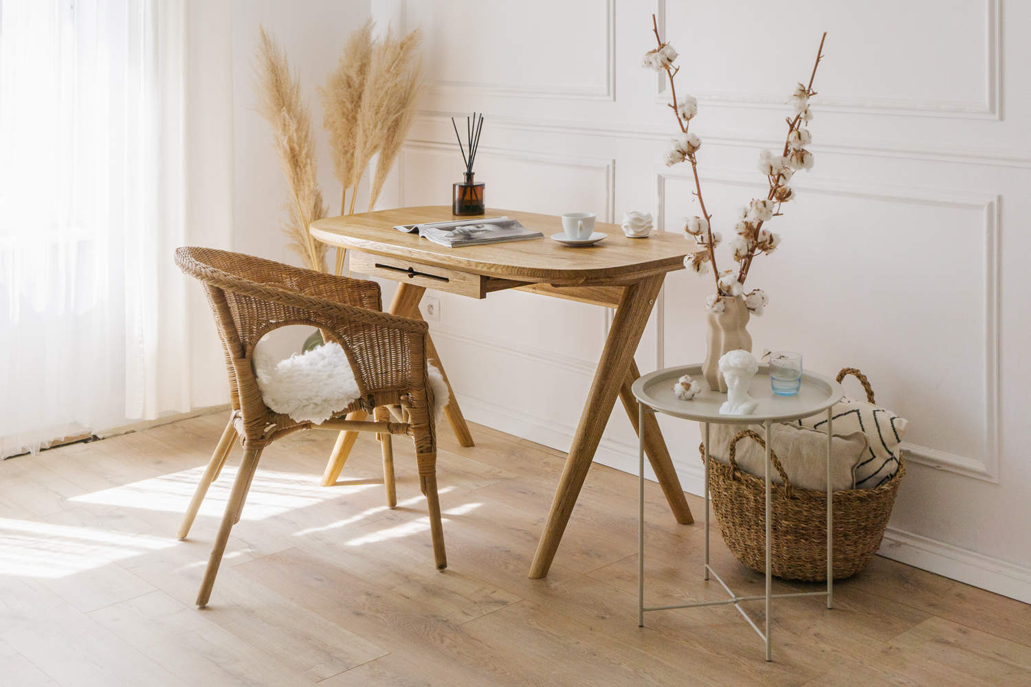 Small solid wood desk for vanity is showcased in a luxury living room with wicker chair and decorative items in a bright room with elegant mouldings. Magazine and cup of coffee laying on rounded corners desk top. 