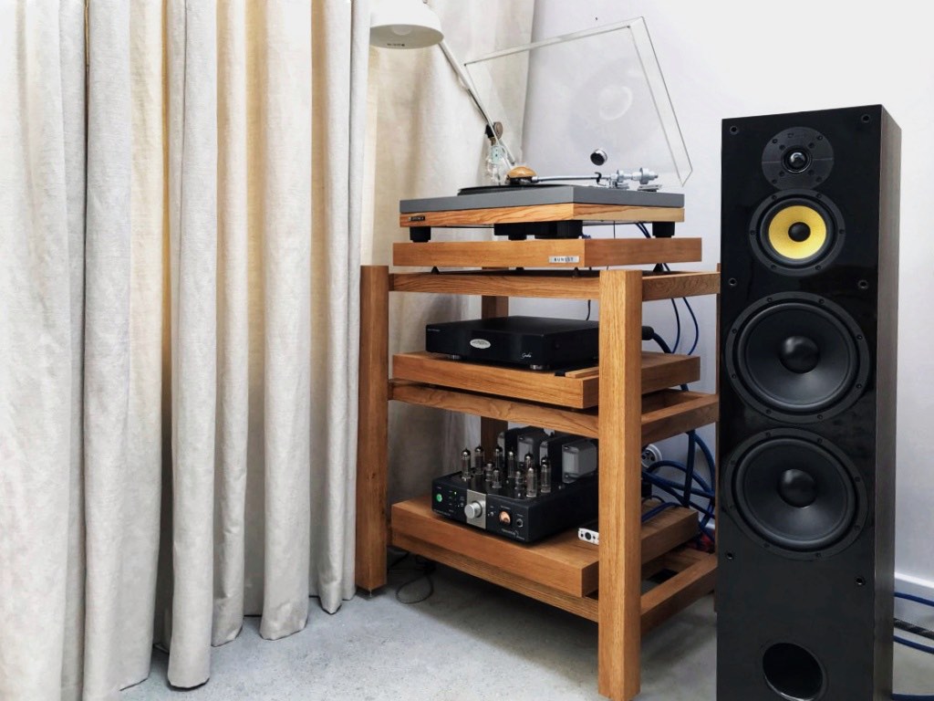 A three-tier home audio rack in a living room corner by a soft curtain, with a Victrola Montauk turntable on top, a Fezz Audio Gratia phono preamp on the middle shelf and a Tsakiridis Hermes tube amplifier below