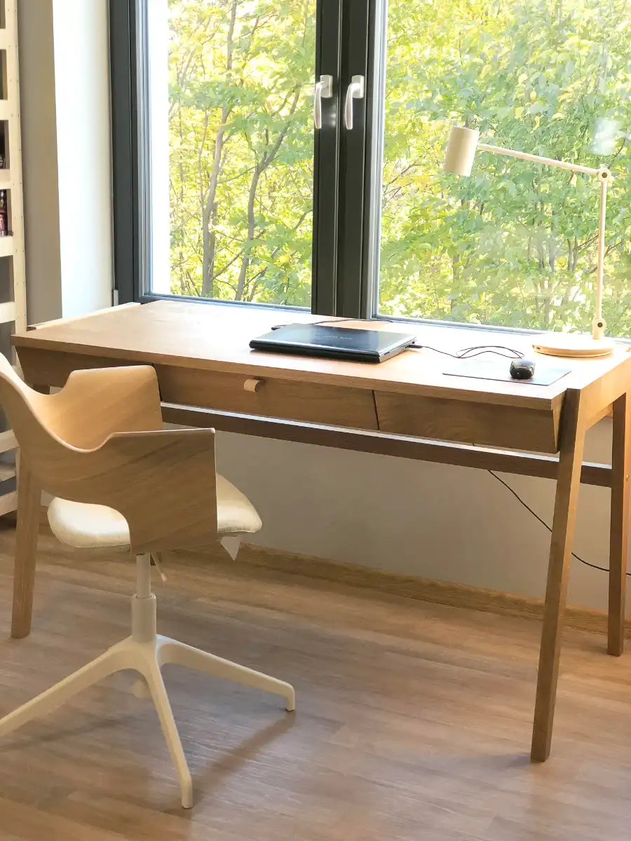 Handmade wooden desk with drawer standing on oak floor in front of the window.
